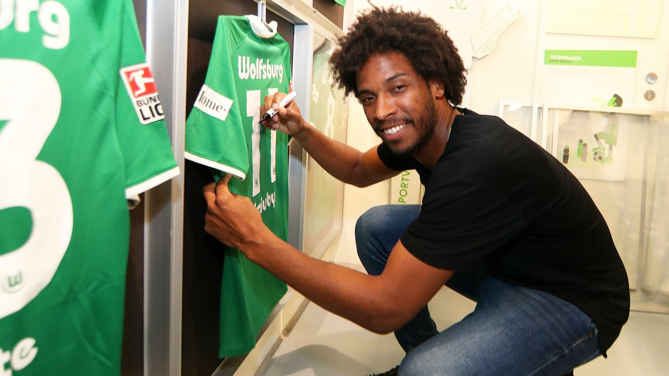 Der ehemalige Spieler des VfL Wolfsburg Caiuby sitzt in der Hocke und unterschreibt auf einem Trikot des VfL Wolfsburg.