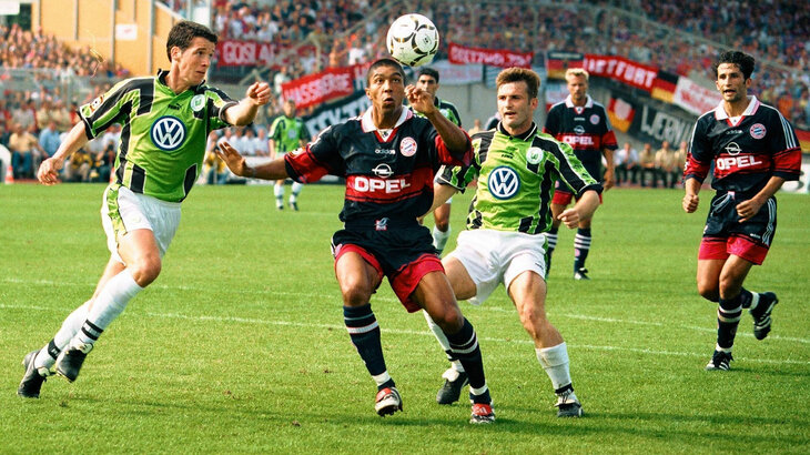 Ehemaliger VfL-Wolfsburg-Spieler Brian O’Neil im Spiel gegen den FC Bayern München. Ein Bayern Spieler hat den Ball vorm Kopf. O’Neil läuft von links zum Ball. Ein weiterer VfL Spieler kommt von rechts.
