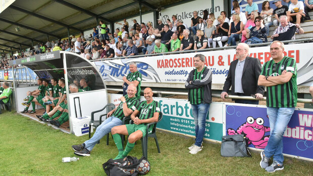 Die Aktiven sitzen auf der Trainerbank und die Zuschauer auf der Tribüne beim Jubiläumsspiel des VfL Wolfsburg in Vechelde.
