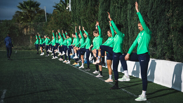 Die VfL-Wolfsburg-Spielerinnen stehen nebeneinander. Alle heben einen Arm und ein Bein hoch.