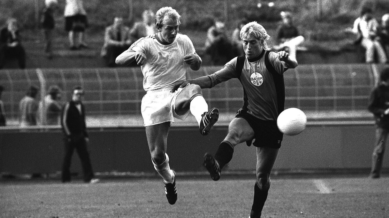 Der ehemalige VfL-Wolfsburg-Spieler Siegfried Otte springt in die Luft und streckt ein Bein zum Ball aus. Neben ihm ist ein Gegenspieler.