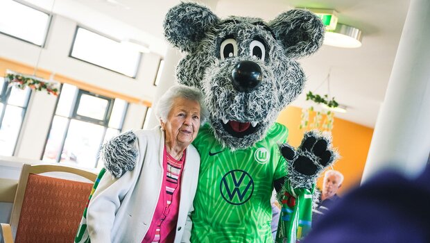 VfL-Wolfsburg-Maskottchen Wölfi und eine AWO-Altenheim-Bewohnerin stehen Arm in Arm.