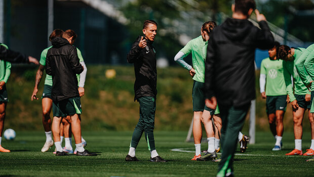 VfL Wolfsburgs Cheftrainer Daniel Bauer steht auf dem Fußballplatz und streckt seine Hand nach vorne. Um ihn stehen die Spieler des VfL.