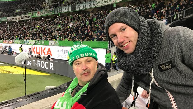 Zwei VfL Wolfsburg-Fans lachen in die Kamera mit der Nordkurve im Hintergrund.