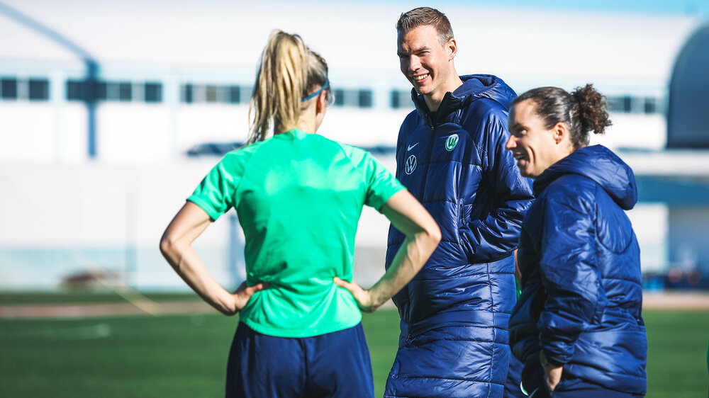 Die VfL-Wolfsburg-Trainer Tommy Stroot und Sabrina Eckhoff stehen lachend bei Kathrin Hendrich.T