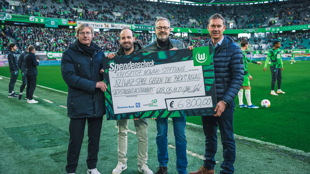 Geschäftsführer des VfL Wolfsburg, Tim Schumacher, nimmt am Spielfeldrand einen Spendenscheck in Empfang.
