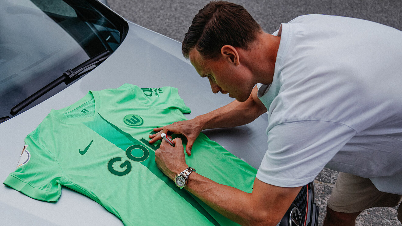 Yannick Gerhardt unterschreibt ein VfL-Wolfsburg-Trikot auf der Motorhaube eines Golfs.