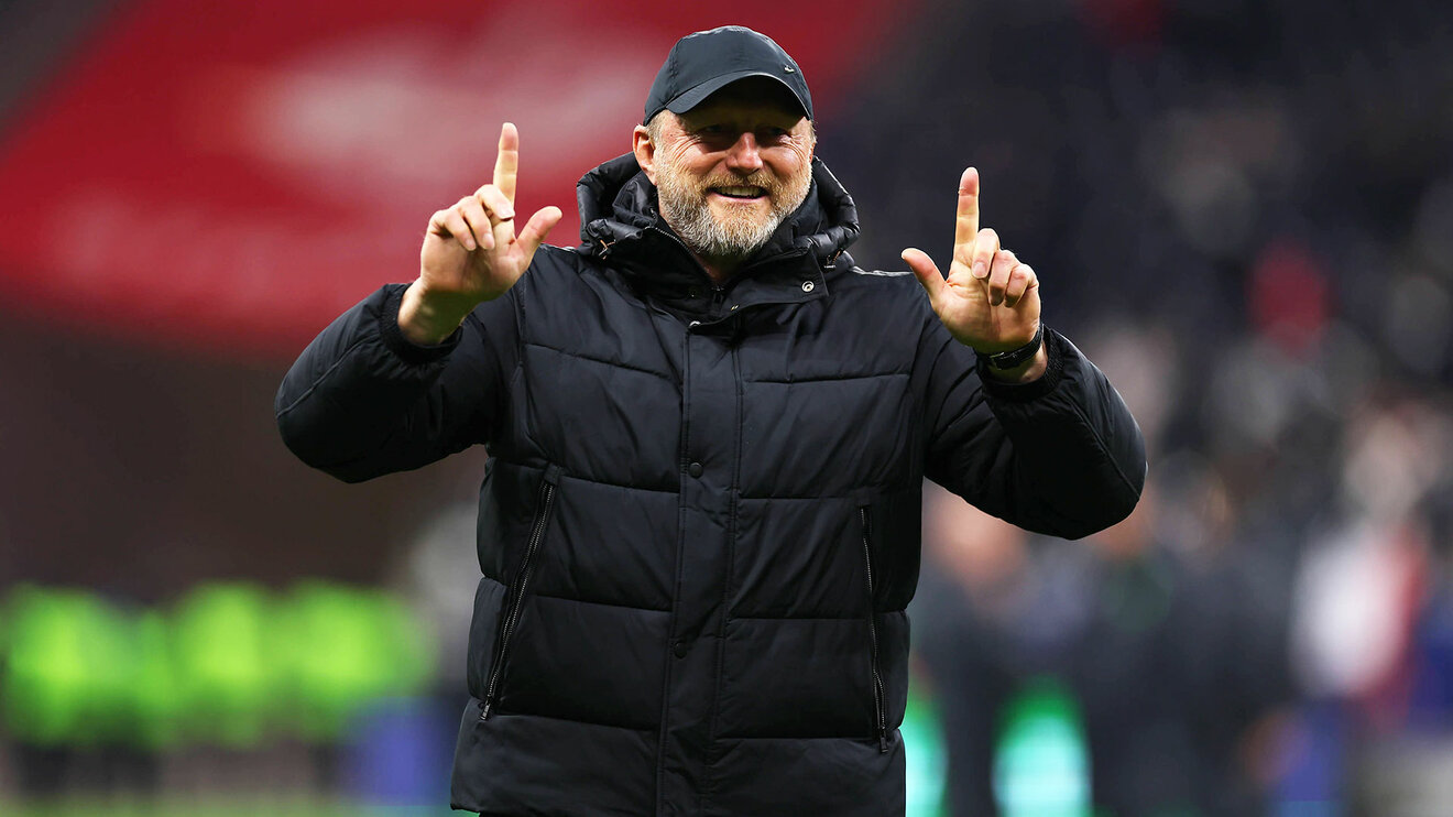 VfL-Wolfsburg-Trainer Ralph Hasenhüttl lächelt und streckt beide Hände nach oben.