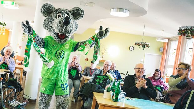 VfL-Wolfsburg-Maskottchen Wölfi steht im Speiseraum des AWO-Altenheim inmitten der Bewohnern und Bewohnerinnen.