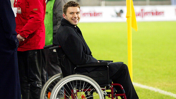 Der ehemalige VfL Wolfsburg Spieler Krzysztof Nowak sitzt im Rollstuhl am Spielfeldrand und lächelt in die Kamera.