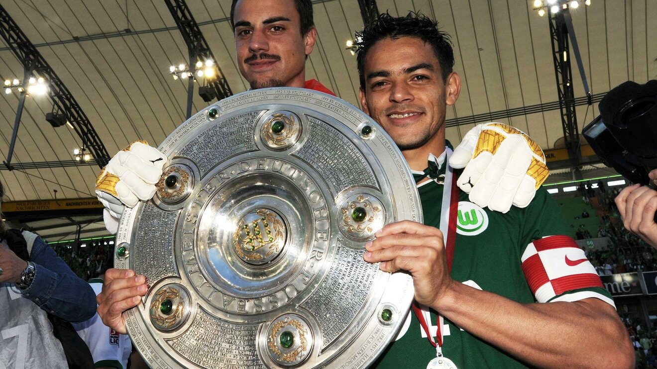 Der ehemalige Spieler Josue des vfL Wolfsburg hält die Meisterschaftsschale in der Hand und lächelt in die Kamera.