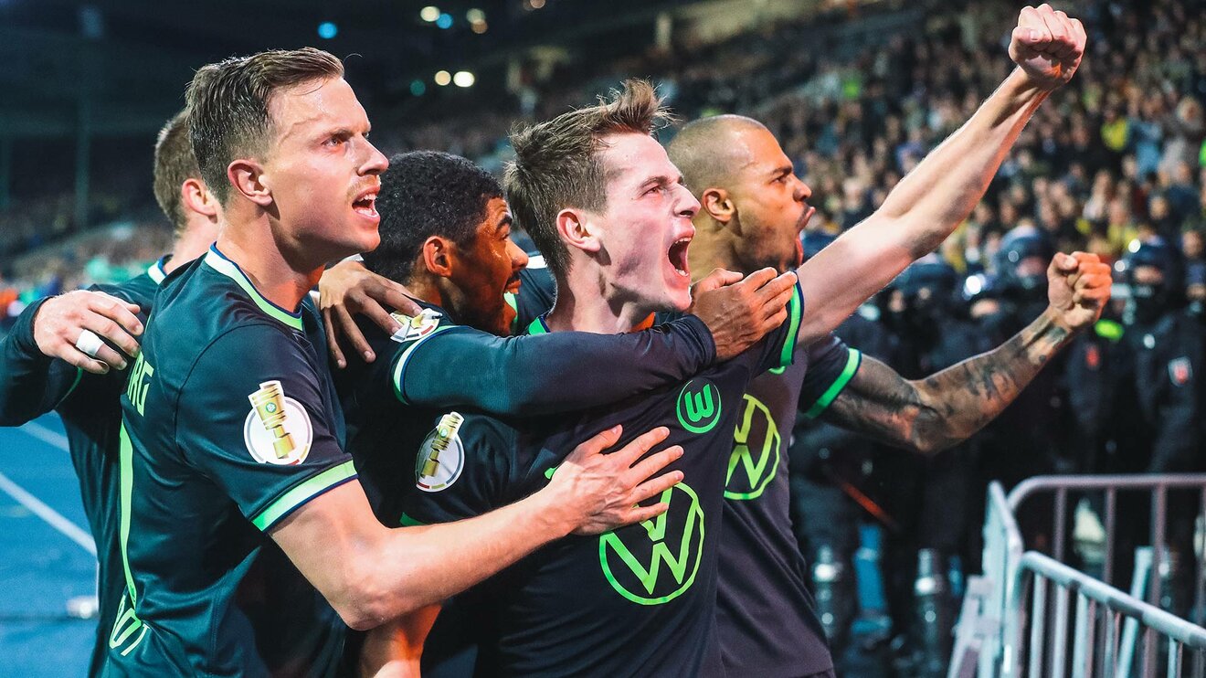 VfL-Wolfsburg-Spieler Jakub Kaminski steht mit Mitspielern vor der Fankurve, streckt seine Faust in die Höhe und jubelt.
