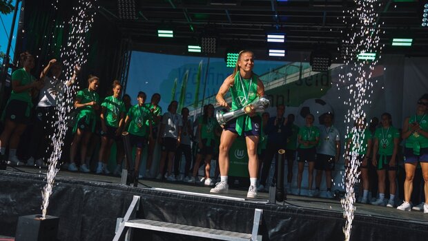 Lynn Wilms vom VfL Wolfsburg hebt auf der Bühne den Pokal in die Luft und jubelt mit ihrem Team.