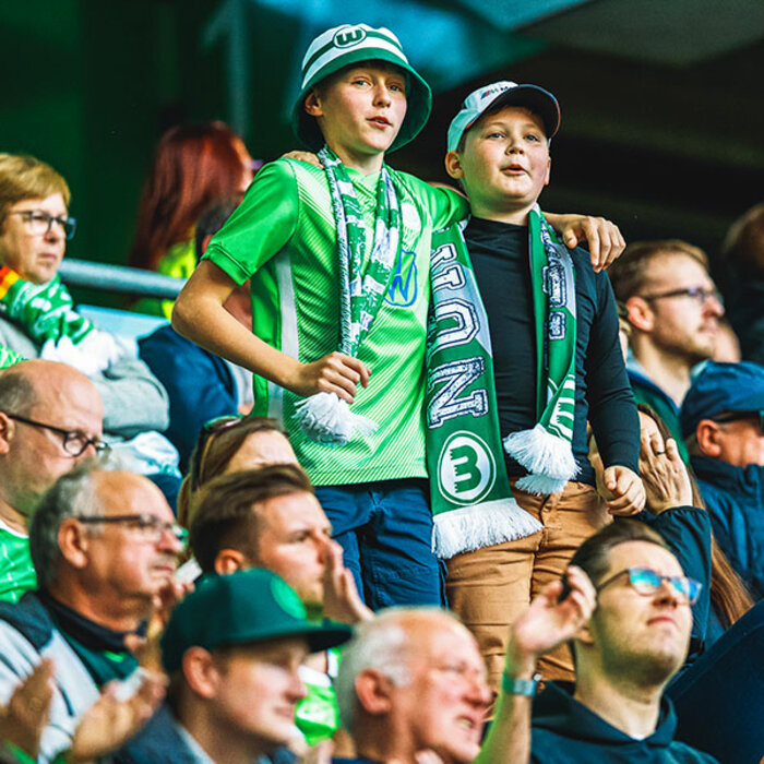 Zwei junge VfL-Wolfsburg-Fans gemeinsam Arm in Arm auf der Tribüne.