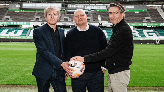Die Geschäftsführung des VfL Wolfsburg steht gemeinsam in der Volkswagen Arena und legt die Hände auf einen Fußball.