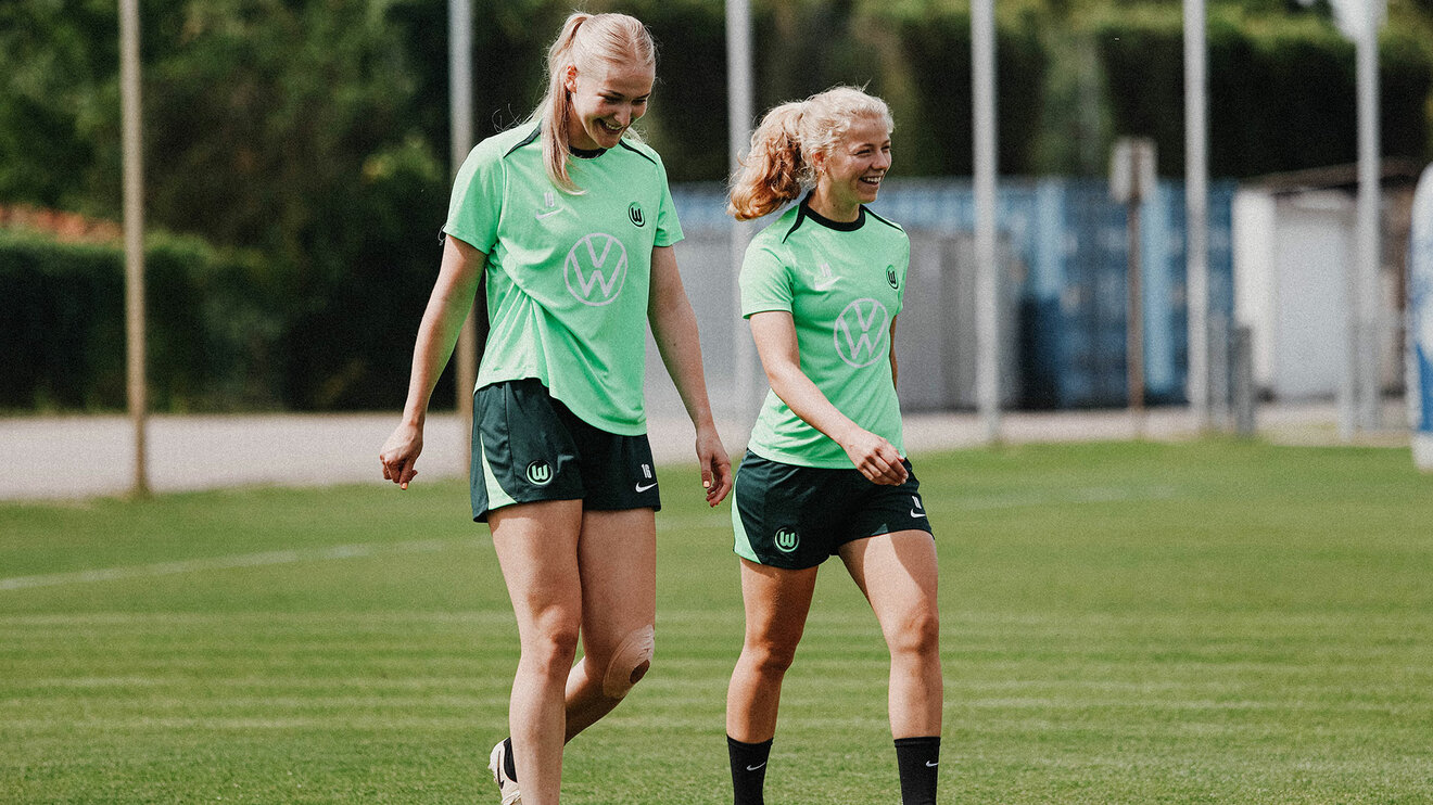 VfL-Wolfsburg-Spielerinnen Camilla Küver und Justine Kielland laufen nebeneinander und lachen.