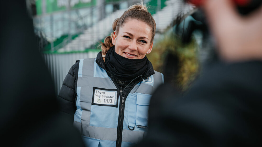 Eine Ordnerin des VfL Wolfsburg lächelt und berät Fans im Stadion.