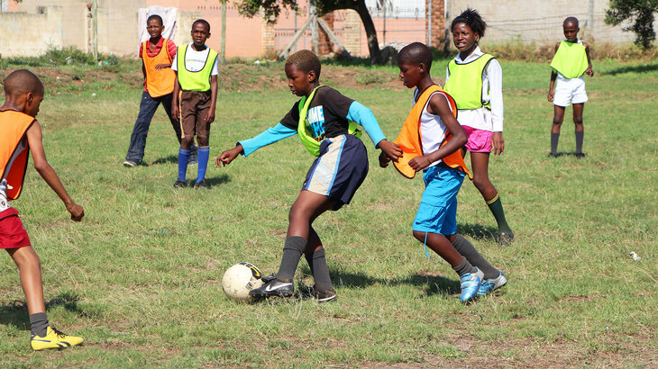 Auf dem Bild sind Kinder zu sehen, welche mithilfe von Leibchen, in Mannschaften eingeteilt sind und miteinander Fußball spielen.
