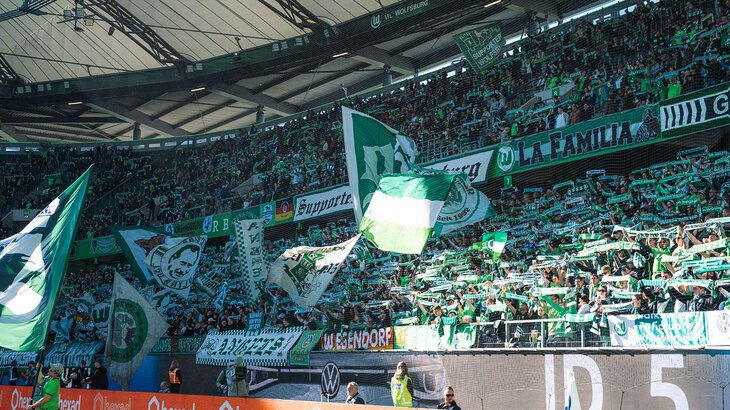 Die Fans des VfL Wolfsburg heben alle ihre Schals in der Fankurve in die Höhe.