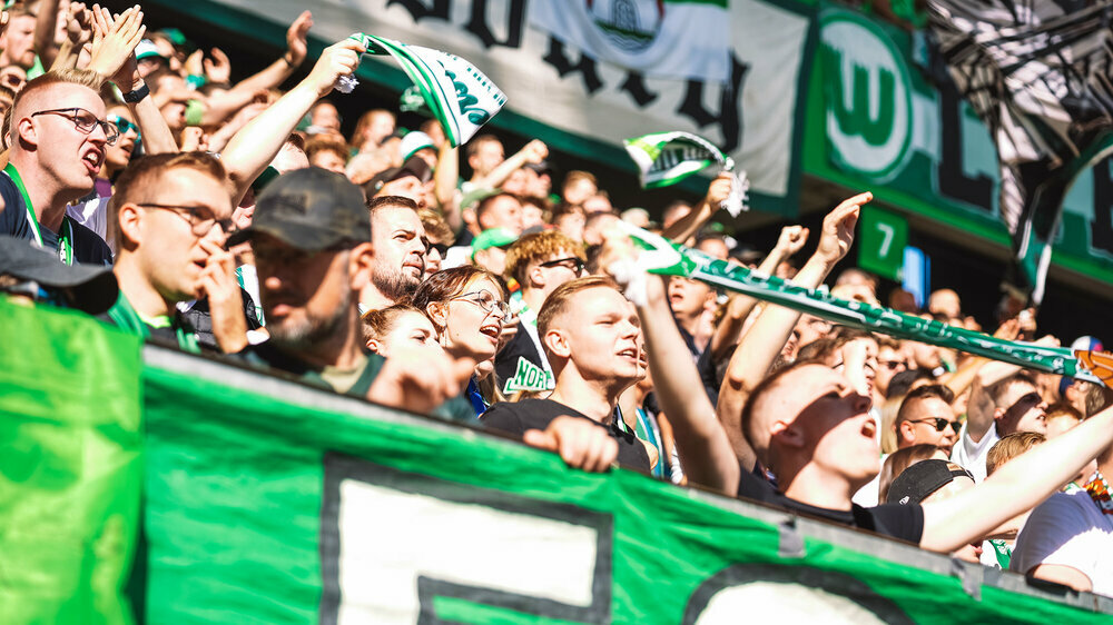 Die Fans des VfL Wolfsburg stehen auf der Tribüne und halten Fahnen sowie Schals in die Luft.