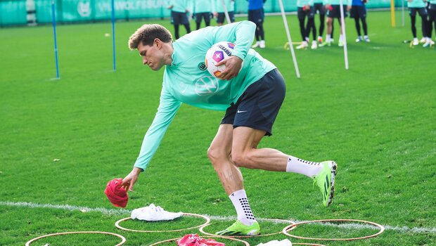 Kevin Behrens vom VfL Wolfsburg hält in einer Hand den Ball und mit der anderen Hand legt er ein Laibchen auf den Boden.