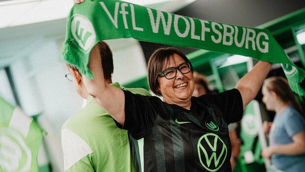Eine Frau posiert im neuen Auswärtstrikot des VfL Wolfsburg und hält einen Fanschal in die Höhe.