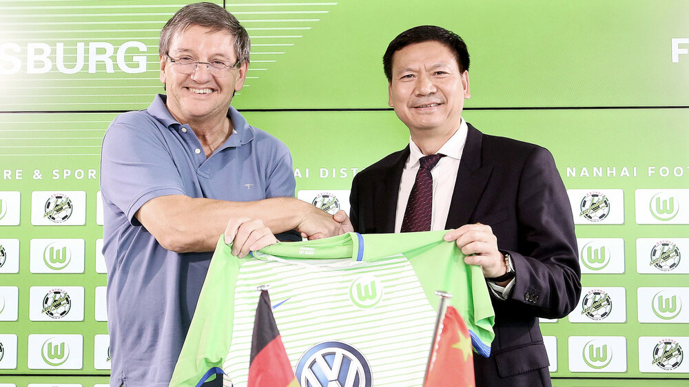 VfL Wolfsburg-Geschäftsführer Wolfgang Hotze und ein chinesischer Partner schütteln sich die Hände und halten ein Trikot hoch.
