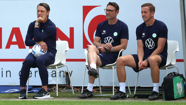 Das VfL Wolfsburg-Trainerteam sitzt auf Stühlen am Spielfeldrand.