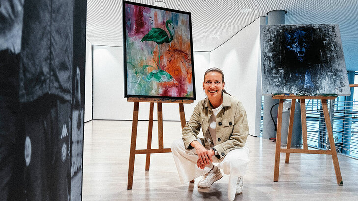 Die Sielerin des VfL Wolfsburg, Joelle Wedemeyer, hockt vor einer Staffelei, auf der ein Kunstwerk zu sehen ist, das einen Flamingo und einen Ball in bunten Farben zeigt.