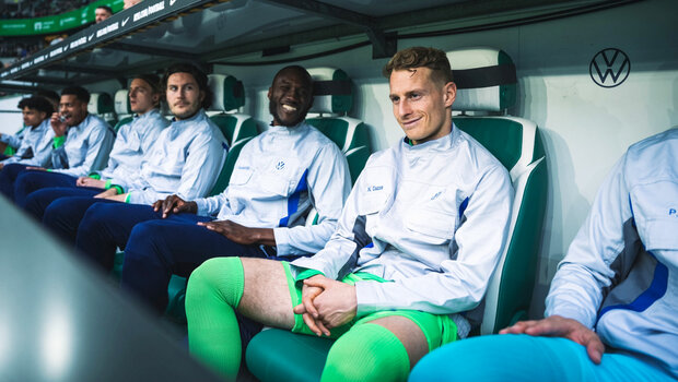 Die VfL-Wolfsburg-Spieler sitzen in den Volkswagen-Arbeitsjacken auf der Ersatzbank.