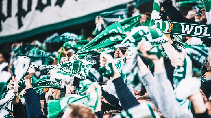 Nahaufnahme von Fans des VfL Wolfsburg, die in der Nordkurve der Volkswagen Arena ihre Schals in die Luft halten.