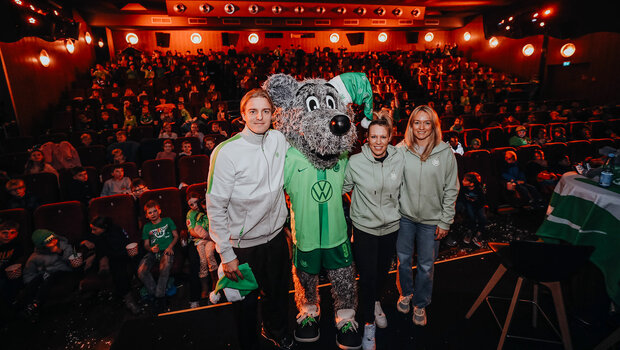 Die VfL-Wolfsburg-Spieler Bornauw, Demann und Schmitz stehen mit Wölfi auf einer Bühne vor Kindern in einem Kinosaal.