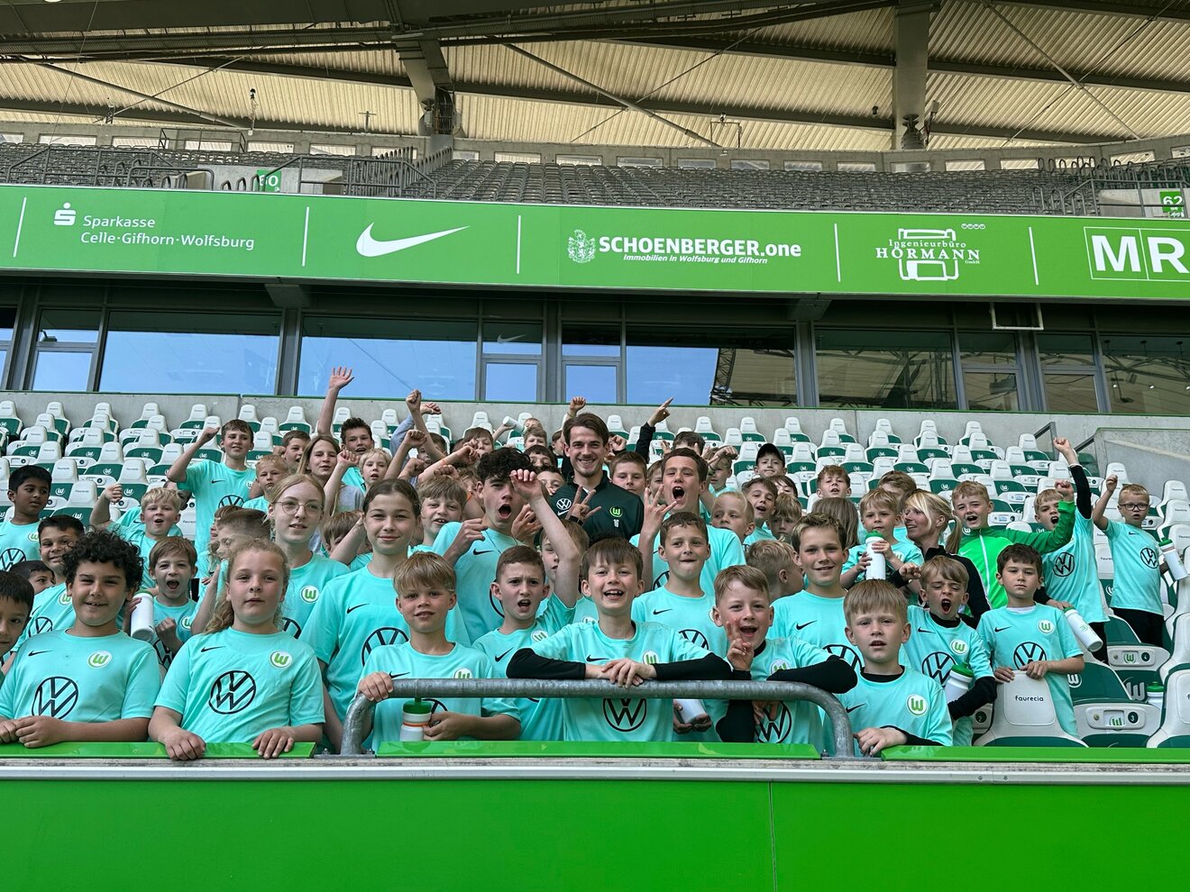 Die Teilnehmer vom VfL Wolfsburg-Osterferiencamp stehen auf der Tribühne und jubeln. In der Mitte von ihnen steht VfL-Profi Jakub Kaminski. 
