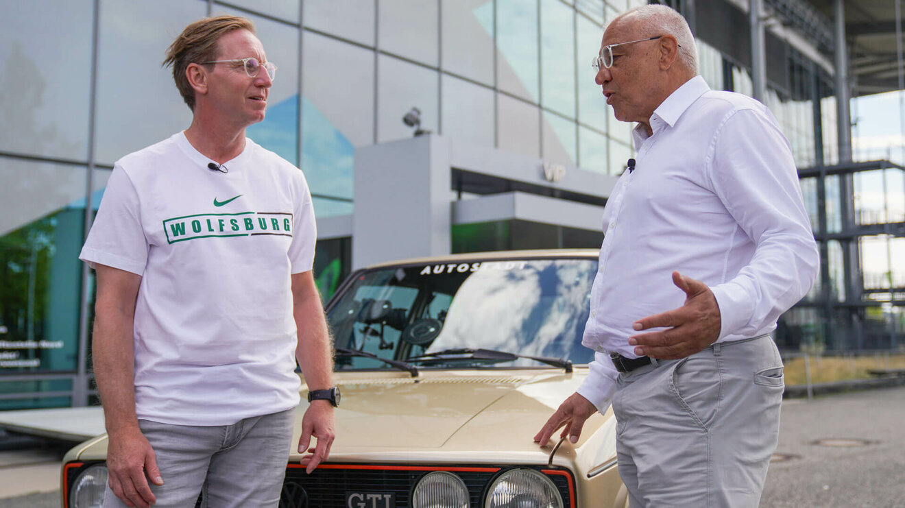 Die VfL-Wolfsburg-Legenden Felix Magath und Roy Präger stehen vor einem alten VW und unterhalten sich.