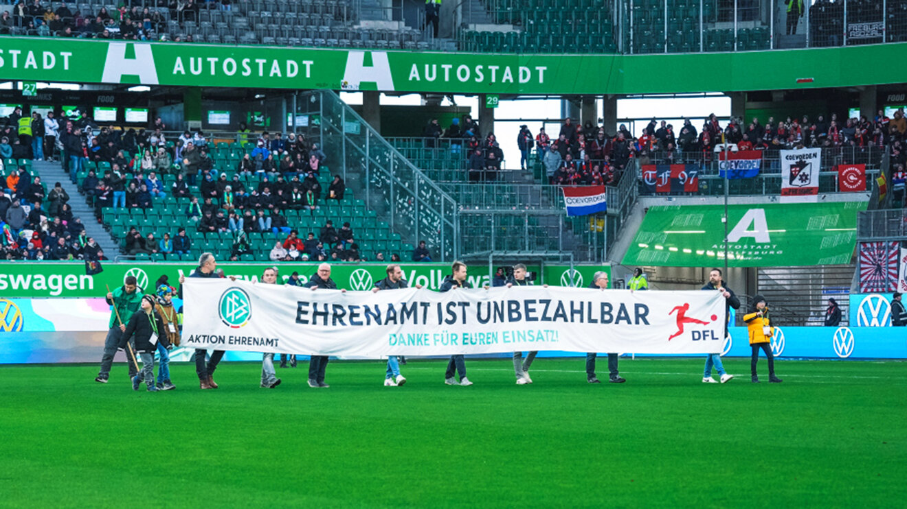 Ehrenamtliche Helfer laufen mit einem Werbebanner für das Ehrenamt über das Spielfeld der Volkswagen Arena.