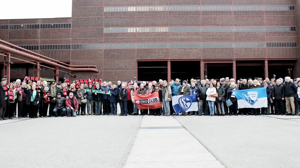 Der Wölfeclub55 Plus des VfL Wolfsburg steht vor einem Ziegelgebäude mit weiteren Seniorengruppen, während sie Banner halten.