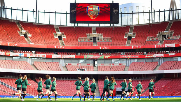 Die Wölfinnen wärmen sich auf dem Platz auf, im Hintergrund die beeindruckende Kulisse der leeren Ränge des Stadions von Arsenal London.