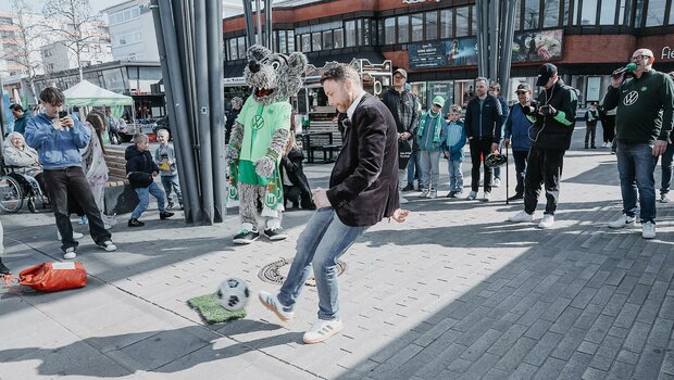 Oberbürgermeister Dennis Weilmann kickt mit ihrem rechten Fuß den Ball in Richtung Torwand. Um Sie herum stehen mehrere Besucher und Wölfi und schauen zu.