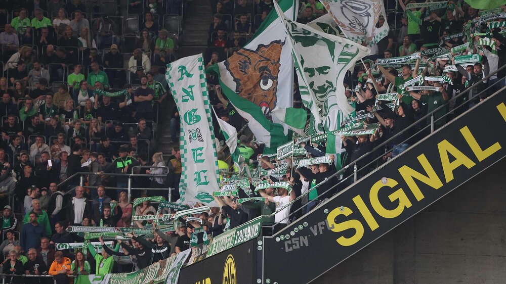 VfL-Wolfsburg-Fans halten Schals und schwenken Fahnen im Oberrang des Signal Iduna Parks.