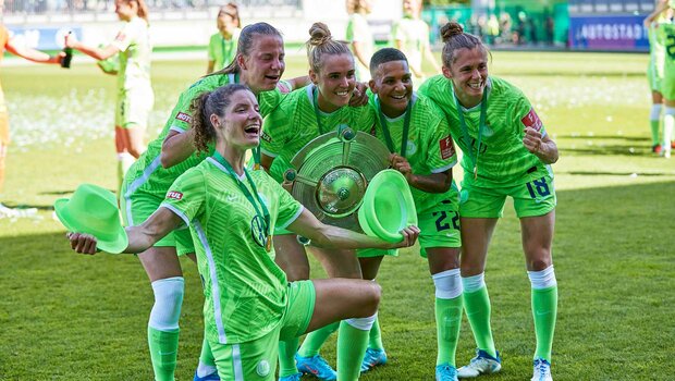 Dominique Janssen vom VfL Wolfsburg hält zwei grüne Hütte 8in den Händen und feiert mit ihren  Mitspielerinnen.