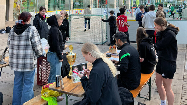 Das Organisationsteam vom VfL Wolfsburg und der Jugendförderung der Stadt Wolfsburg sitzen an einer Bierzeltgarnitur. Darum stehen Mensch und es ist das Spielfeld des Streetkick für Vielfalt zu sehen.