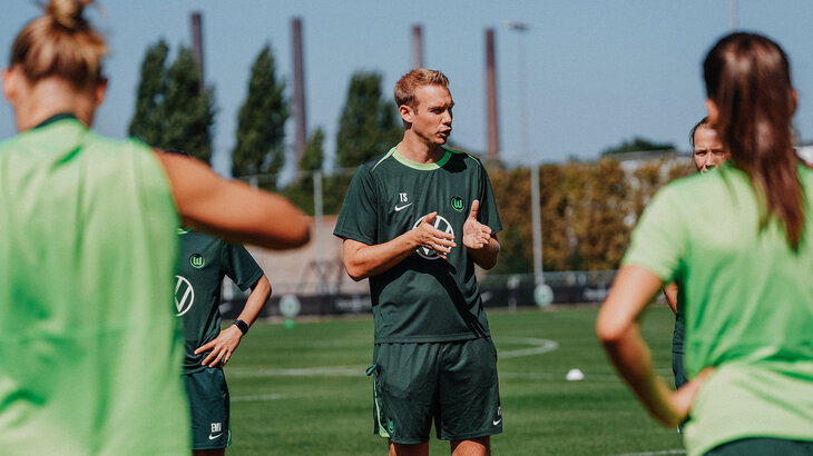 VfL-Wolfsburg-Frauentrainer Tommy Stroot steht inmitten der Spielerinnen und spricht mit ihnen. 