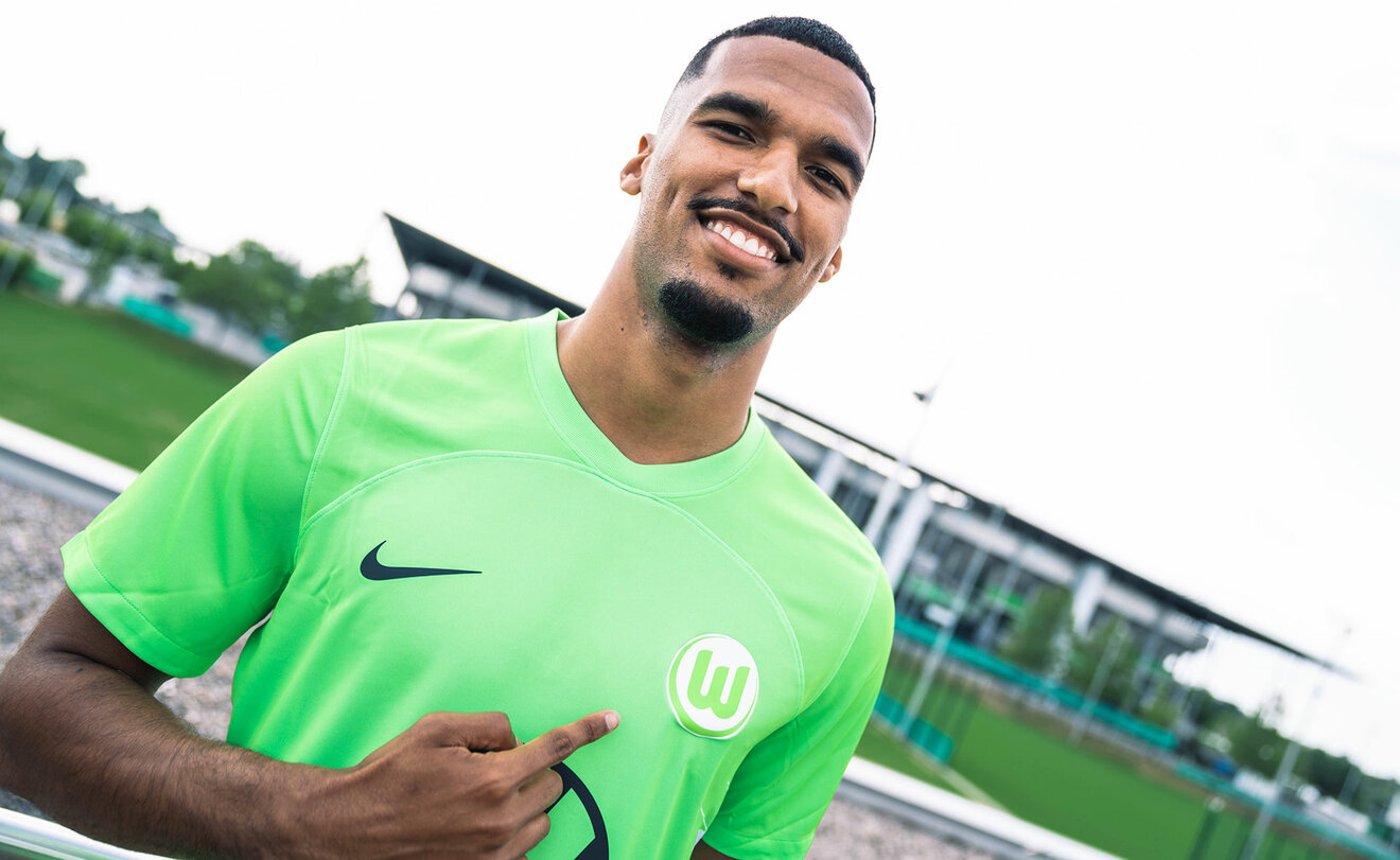 VfL Wolfsburg Neuzugang Moritz Jenz posiert, auf das Logo auf deiner Brust zeigend, im VfL Trikot vor der Volkswagen Arena.
