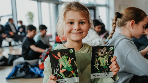 Ein Mädchen hält lachend Autogramme von VfL-Wolfsburg-Spielerin Kristin Demann und Wölfi in den Händen.