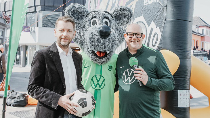 Oberbürgermeister Dennis Weilmann mit Ball in der Hand, daneben Wölfi und ein Moderator vom VfL Wolfsburg.