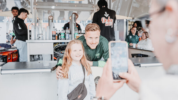 Yannick Gerhardt vom VfL Wolfsburg lehnt sich aus dem Bierwagen. Davor steht ein kleines Mädchen. Eine Frau macht mit ihrem Handy ein Foto von den beiden.