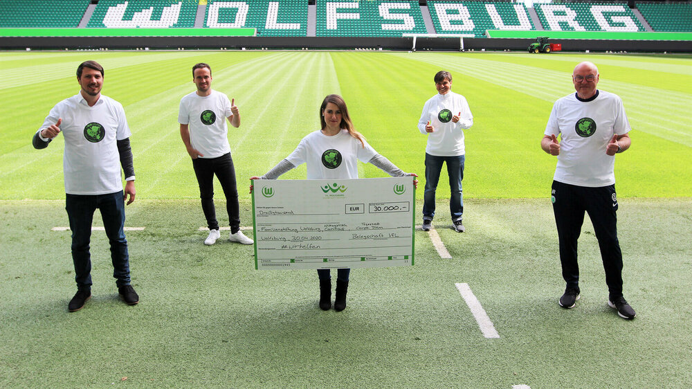 Mitarbeiter des VfL Wolfsburg präsentieren die Spende über 30.000 Euro vor dem Spielfeld der Volkswagen Arena. 