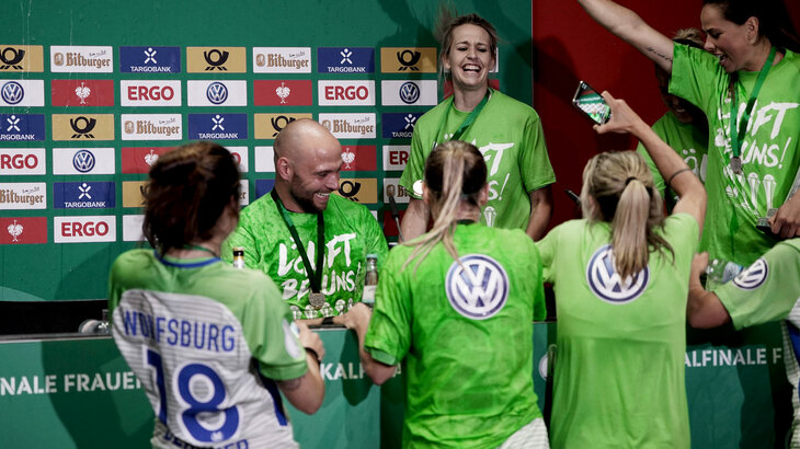 VfL Wolfsburg-Frauentrainer Stephan Lerch wird von einigen feiernden Spielerinnen bei der Pressekonferenz unterbrochen.