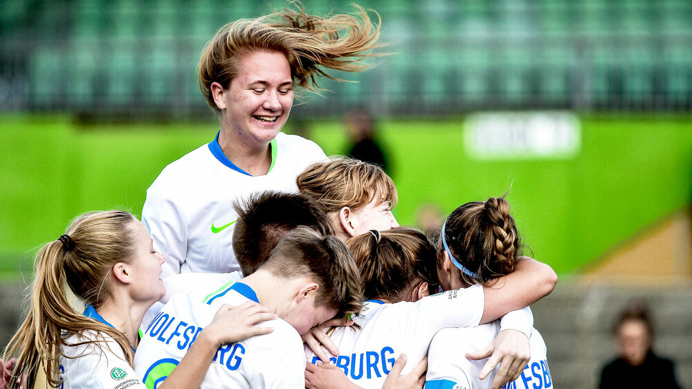 Spielerinnen der U17 Frauenmanschaft des VfL Wolfsburg liegen sich jubelnd im Arm, während eine von ihnen freudig in die Luft springt.