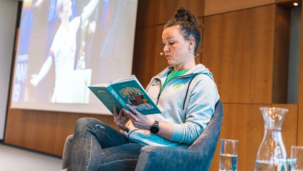 VfL-Wolfsburg-Spielerin Marina Hegering hat ein Buch auf dem Schoß und liest daraus vor.
